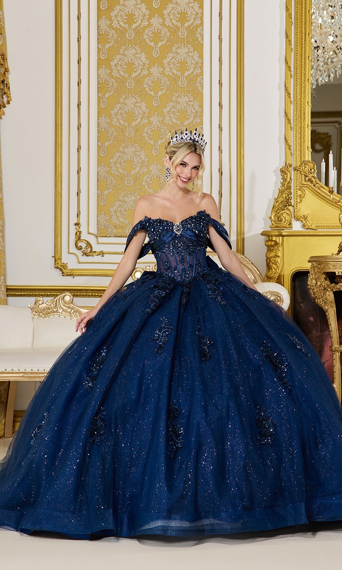A woman wears the Quinceanera Dress 5101 by Dancing Queen in navy, featuring beaded applique, as she stands in an ornate gold-accented room with a cream sofa and a fireplace.
